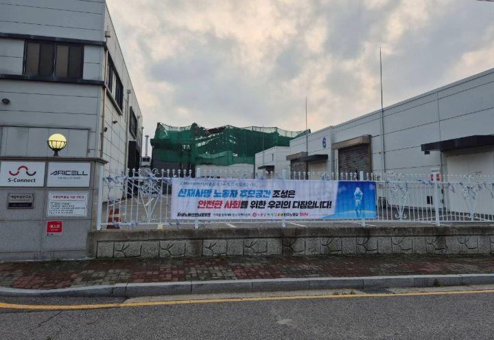 아리셀 참사 2주기를 맞아 걸린 현수막 뒤로 불탄 공장의 모습이 보인다